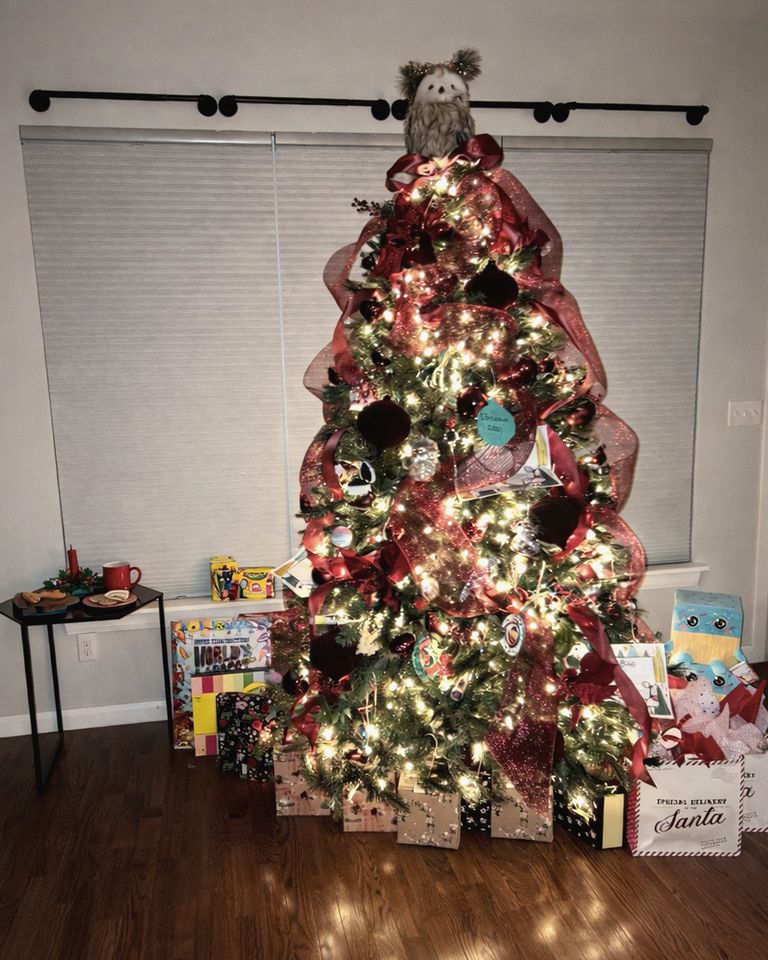 Lit Christmas tree wrapped in red mesh ribbon with wrapped gifts underneath, glowing in the evening